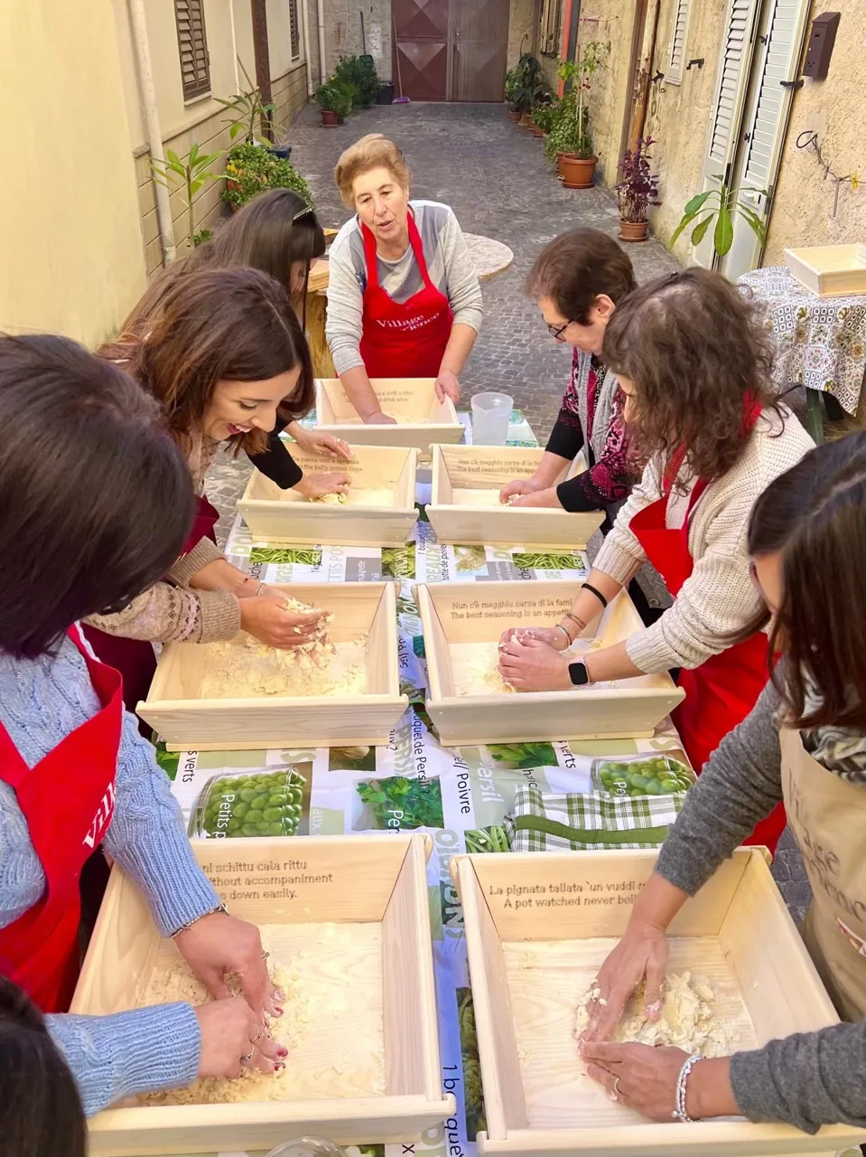 Persone riunite intorno a una tavola in un vicolo preparano insieme un impasto, mentre una donna mostra la preparazione.